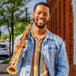A man wearing a jean jacket holding a saxophone smiling.