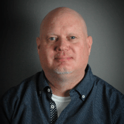 A bald man wearing a grey button up shirt smiling.