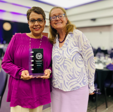 Woman on the left wearing a bright pink shirt holding an award and a woman on the right wearing a light purple patterned top both smiling. 