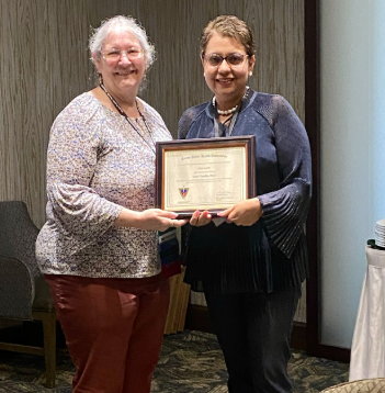 Woman on the left wearing a white and purple floral shirt with brown pants and a woman on the right wearing a blue long sleeve shirt wearing dark pants. 