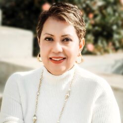 A woman wearing a white sweater with a long necklace and short brown hair smiling.