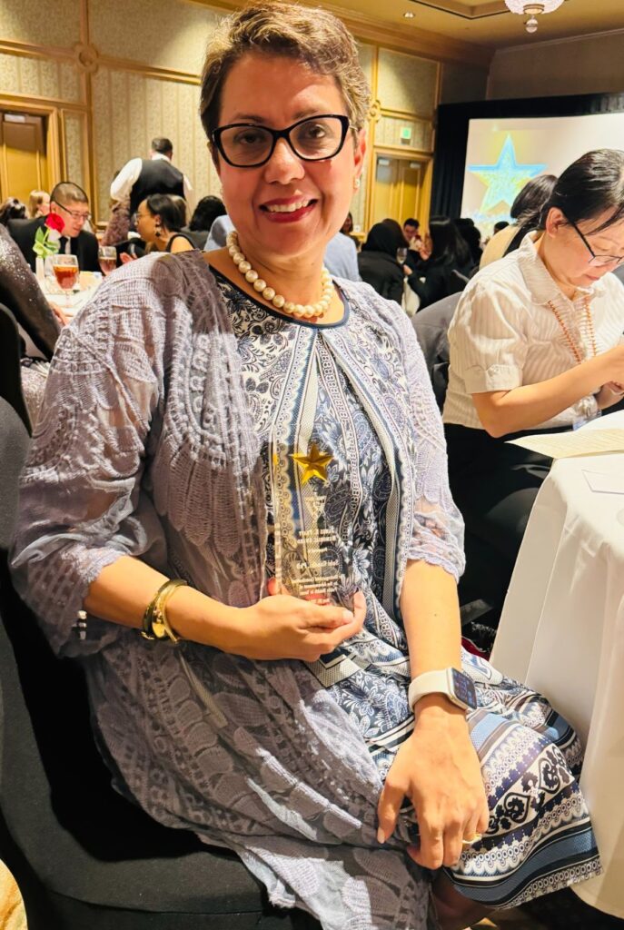 A woman wearing a blue printed dress with short hair and a peal necklace smiling. 