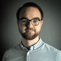 A man wearing a grey stripped button down with a brown beard smiling.