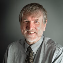 A man wearing a grey shirt with a brown tie is smiling.