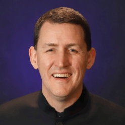 A man wearing a black button up shirting with short brown hair smiling.