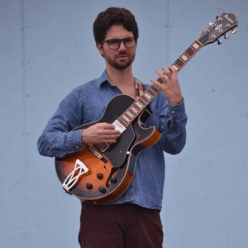 A man with a jean shirt and purple pants playing a brown guitar.