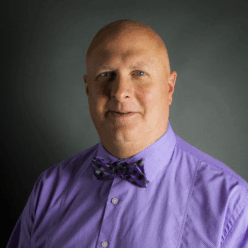 A bald man wearing a purple button up shirt with a plaid purple bowtie.